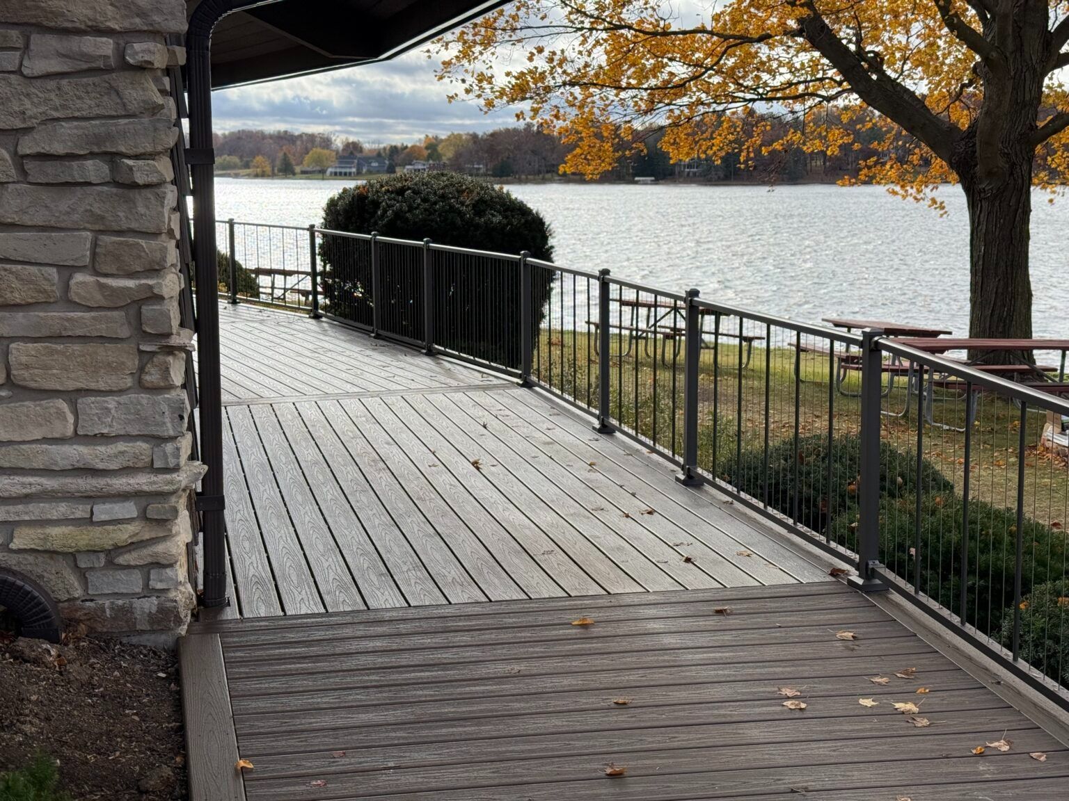 Wooden lakeside deck with black railing, stone wall, and autumn trees overlooking calm water