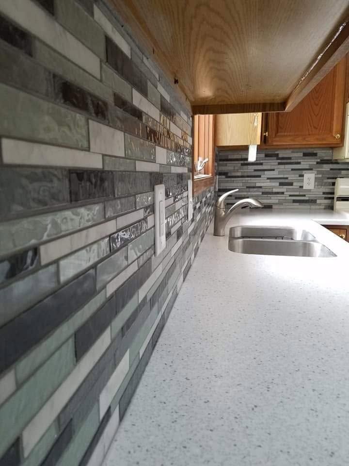 Narrow kitchen with gray mosaic backsplash, white countertop, sink, and wood cabinets.