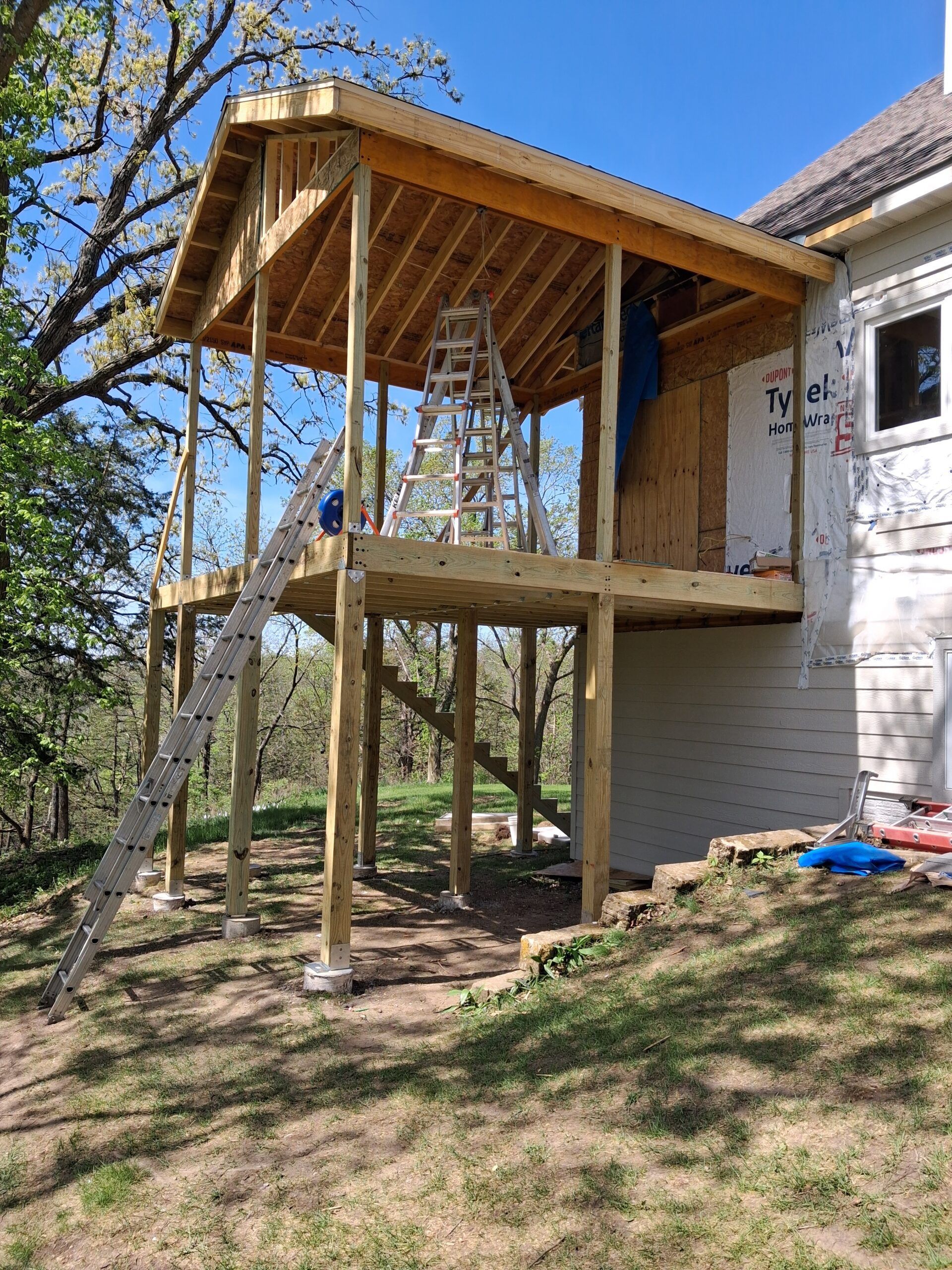 Partially built wooden porch deck attached to a house, with exposed beams and support posts outdoors.