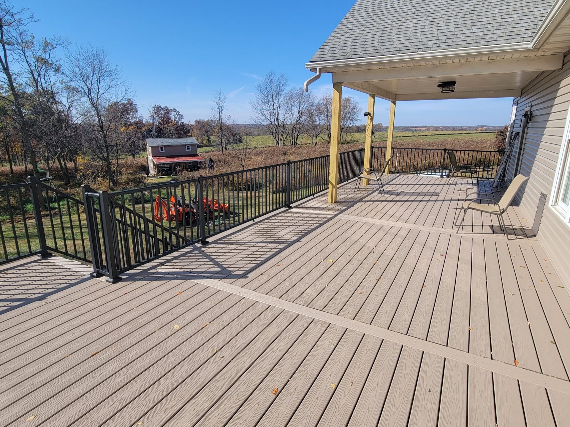 Spacious covered porch with tan decking, black railing, and view of a rural field and red barn.
