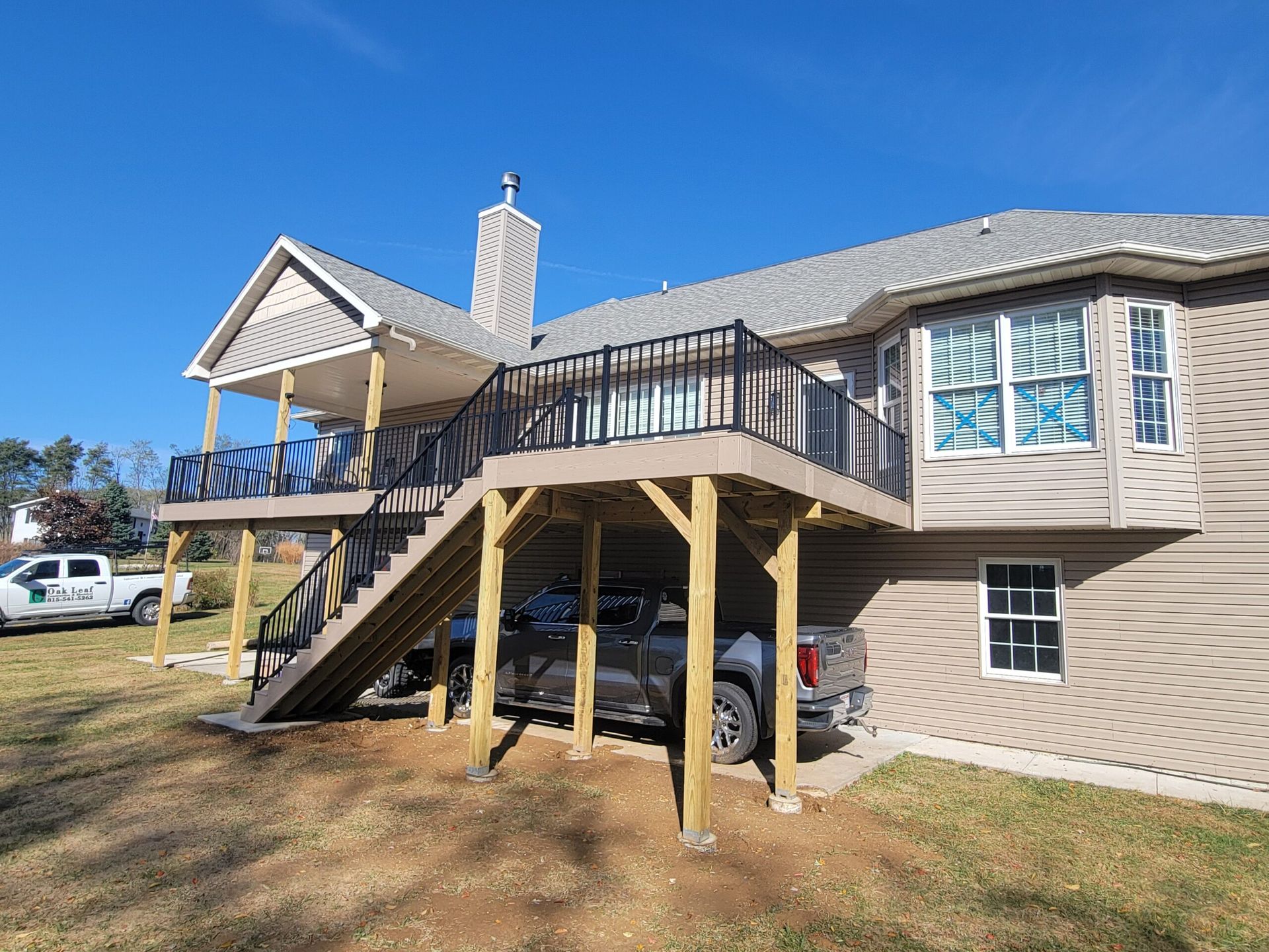 House with elevated wooden deck and staircase, white siding, blue sky, and parked vehicles below.