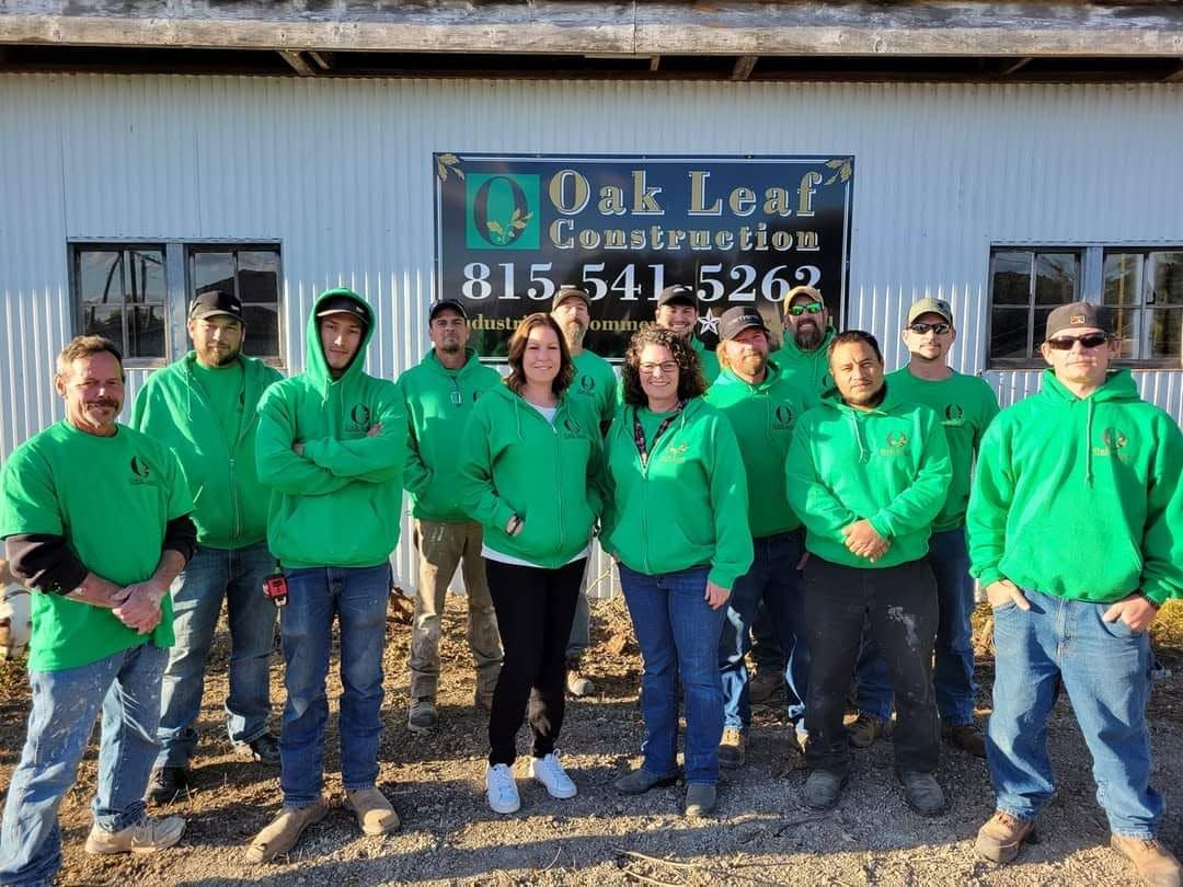 Ten people in green hoodies stand outside an Oak Leaf Construction building sign.