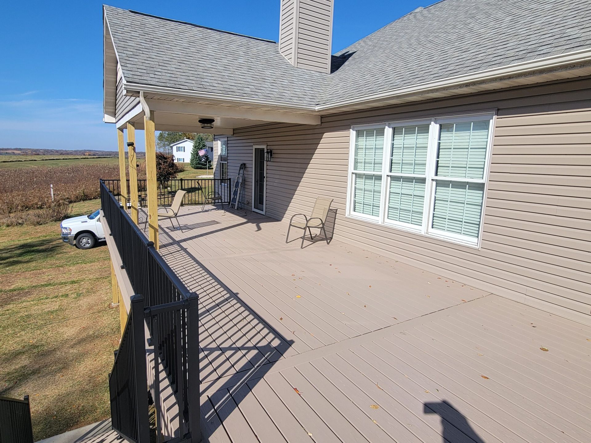 Front porch of a beige house with a wooden deck, black railing, and windows overlooking open fields.