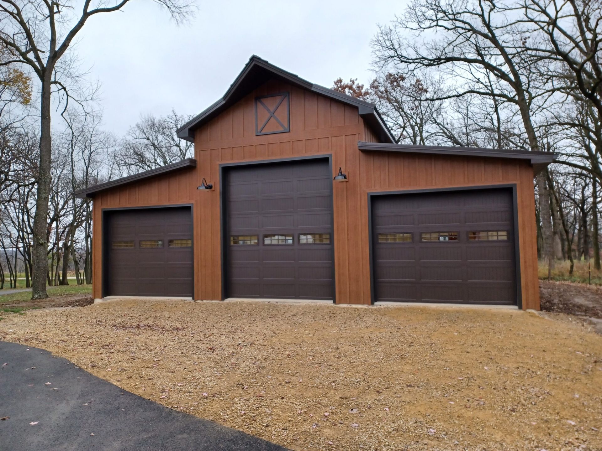 Three-car brown garage with barn-style doors on a wooded gravel lot