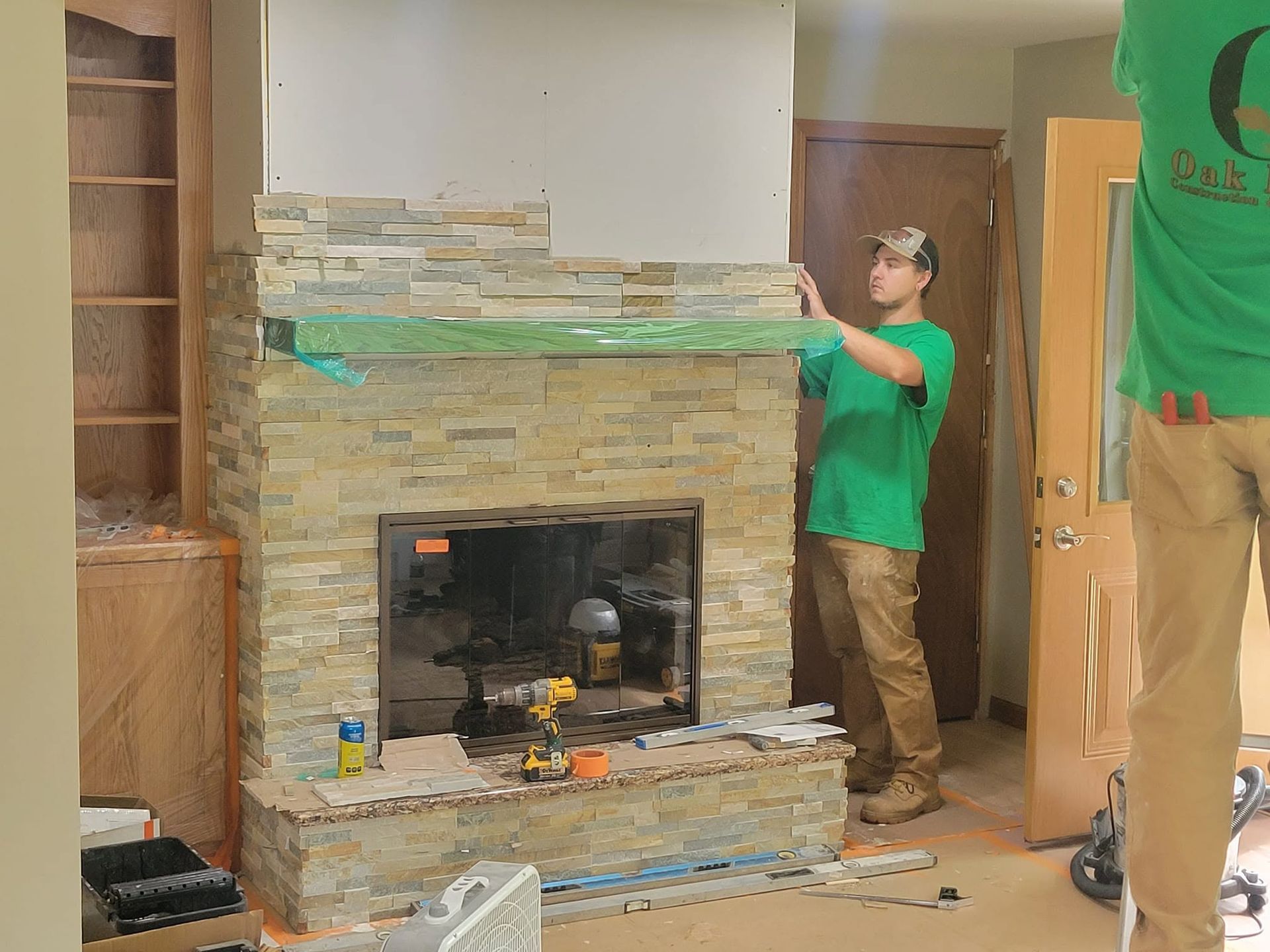 Two workers in green shirts installing stone veneer around a fireplace in a room under renovation