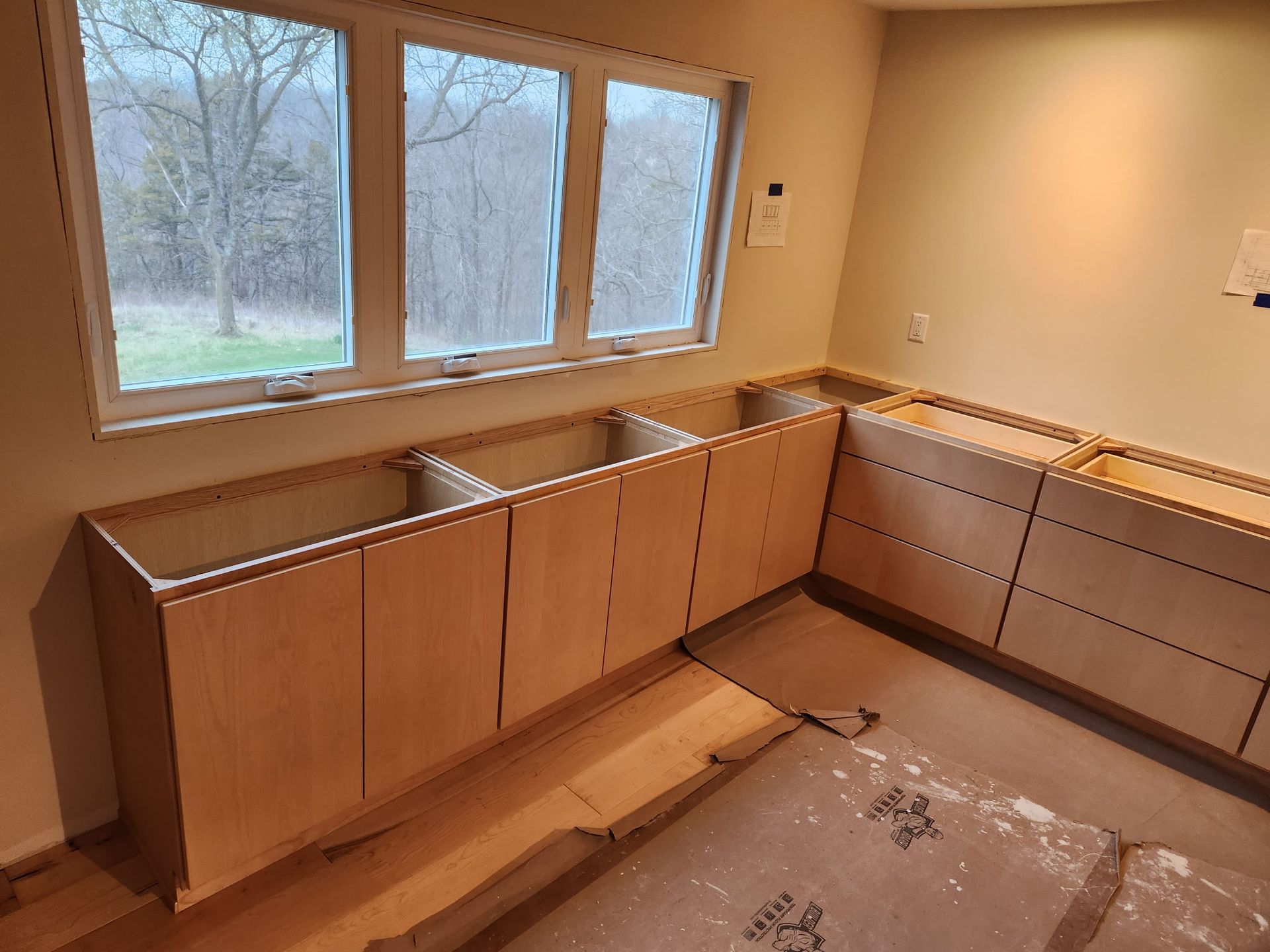 Unfinished kitchen with wood cabinets and countertop under a large window, floor under renovation.