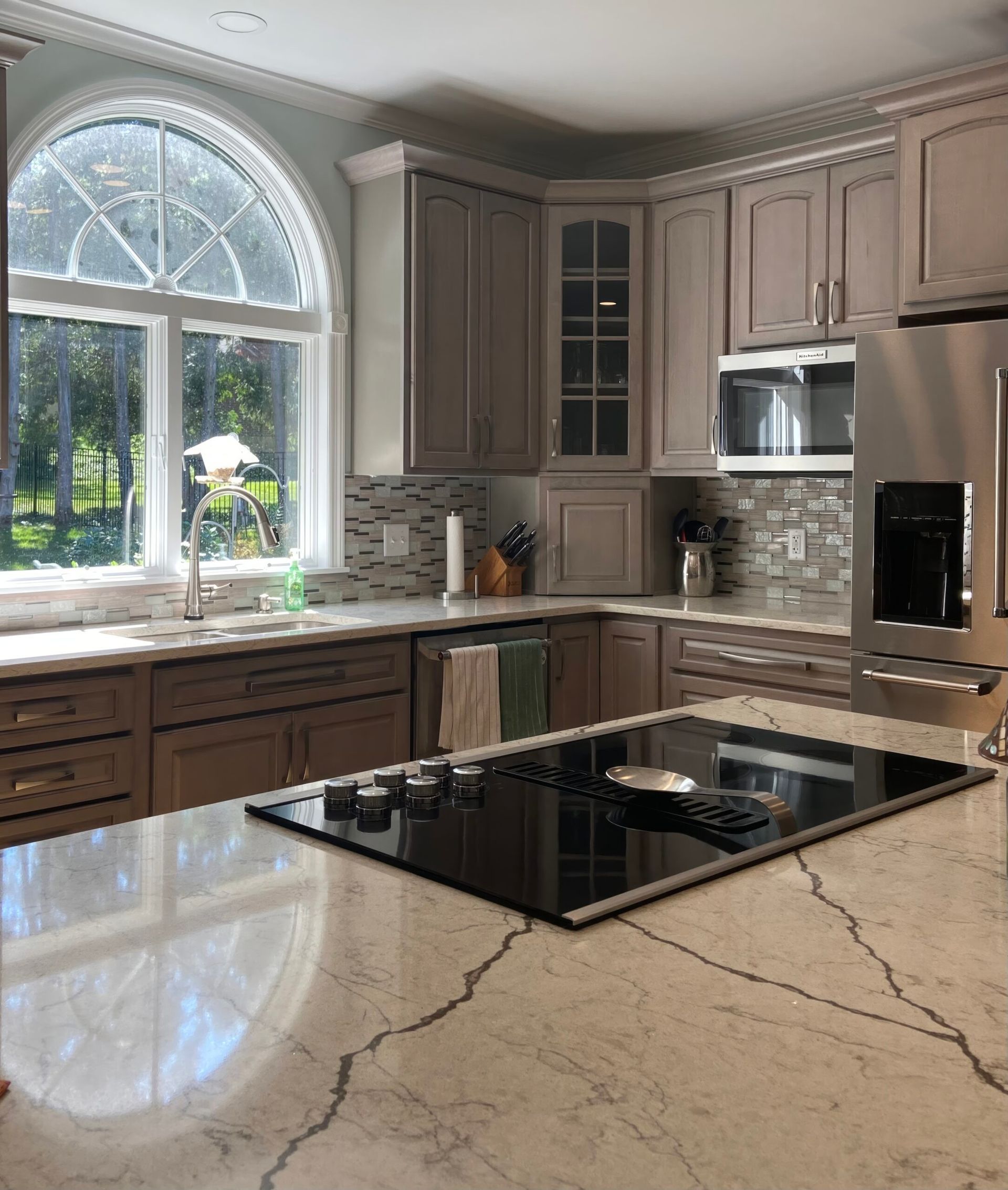 Modern kitchen with light wood cabinets, large arched window, and black island cooktop.