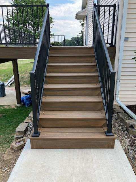 Outdoor staircase with tan steps, black railings, and a concrete landing beside a house.