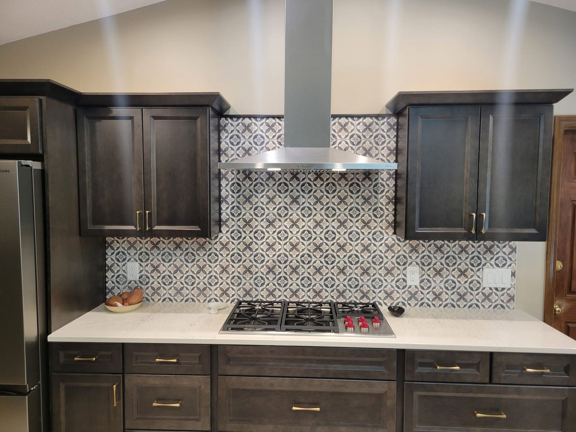Modern kitchen with dark cabinets, patterned tile backsplash, white countertops, and a stainless range hood