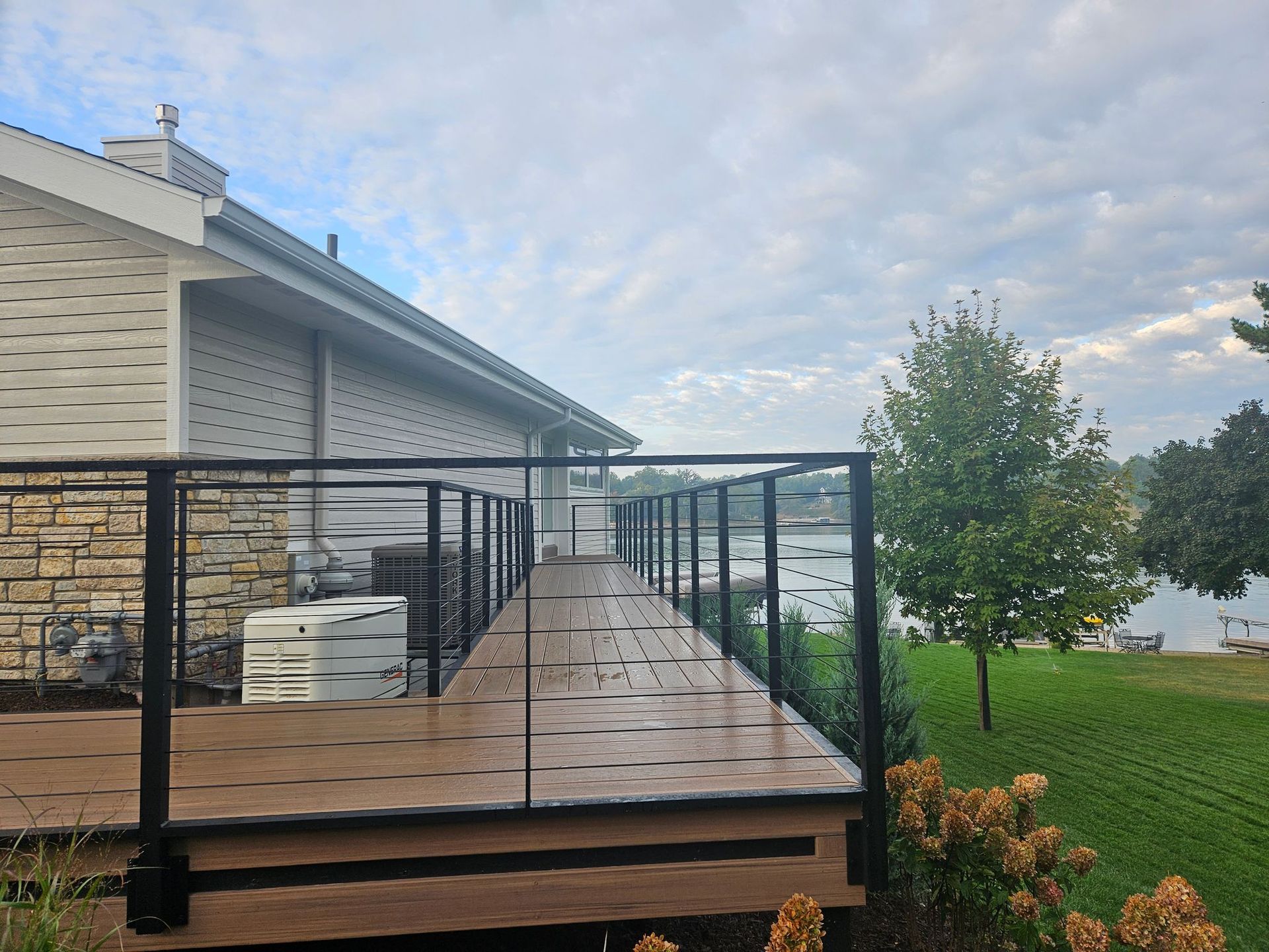 Wooden deck with black railings beside a house, overlooking a grassy yard and lake beyond.