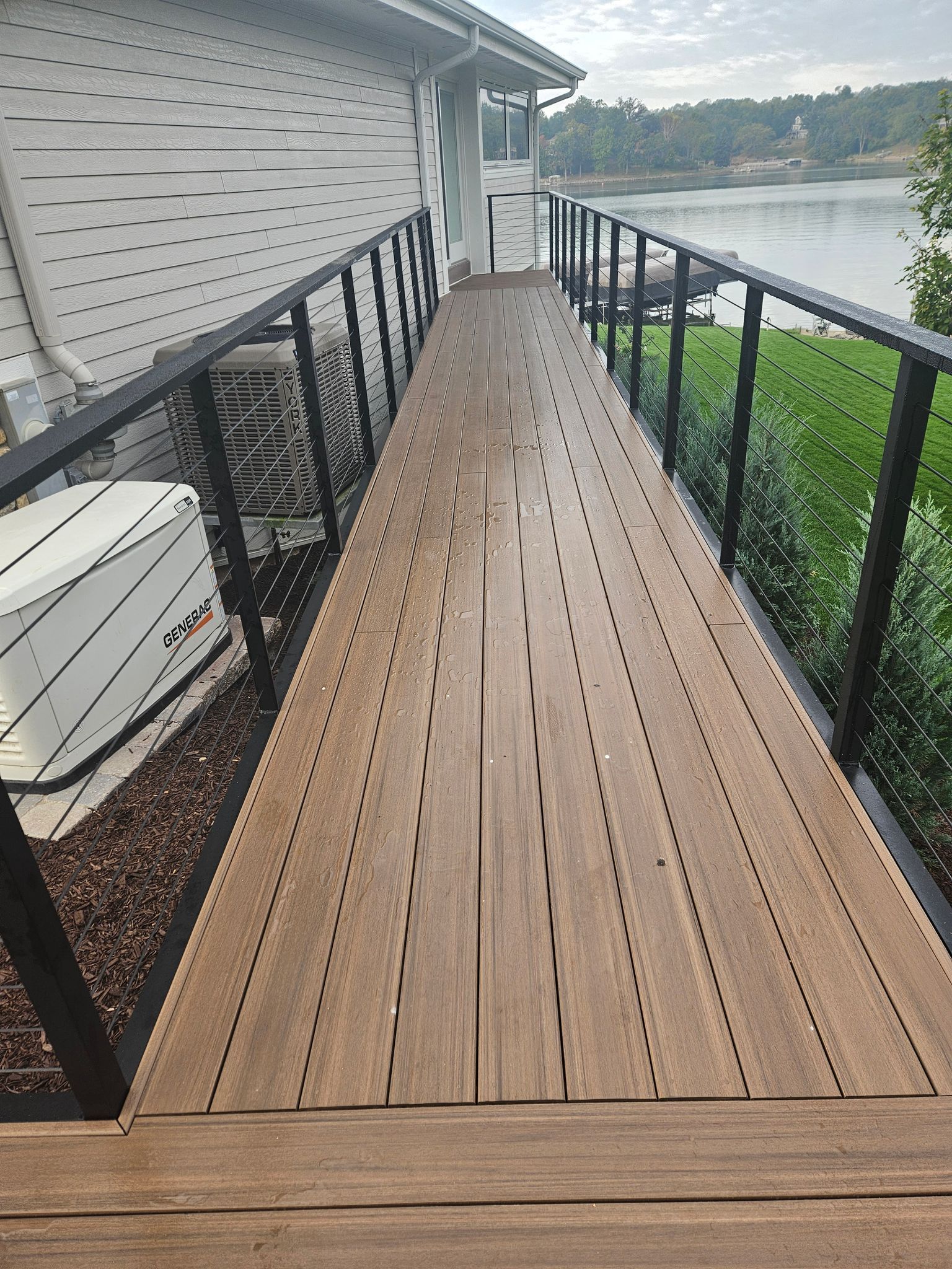 Narrow wooden deck walkway beside a house, with black railings and a lake view.