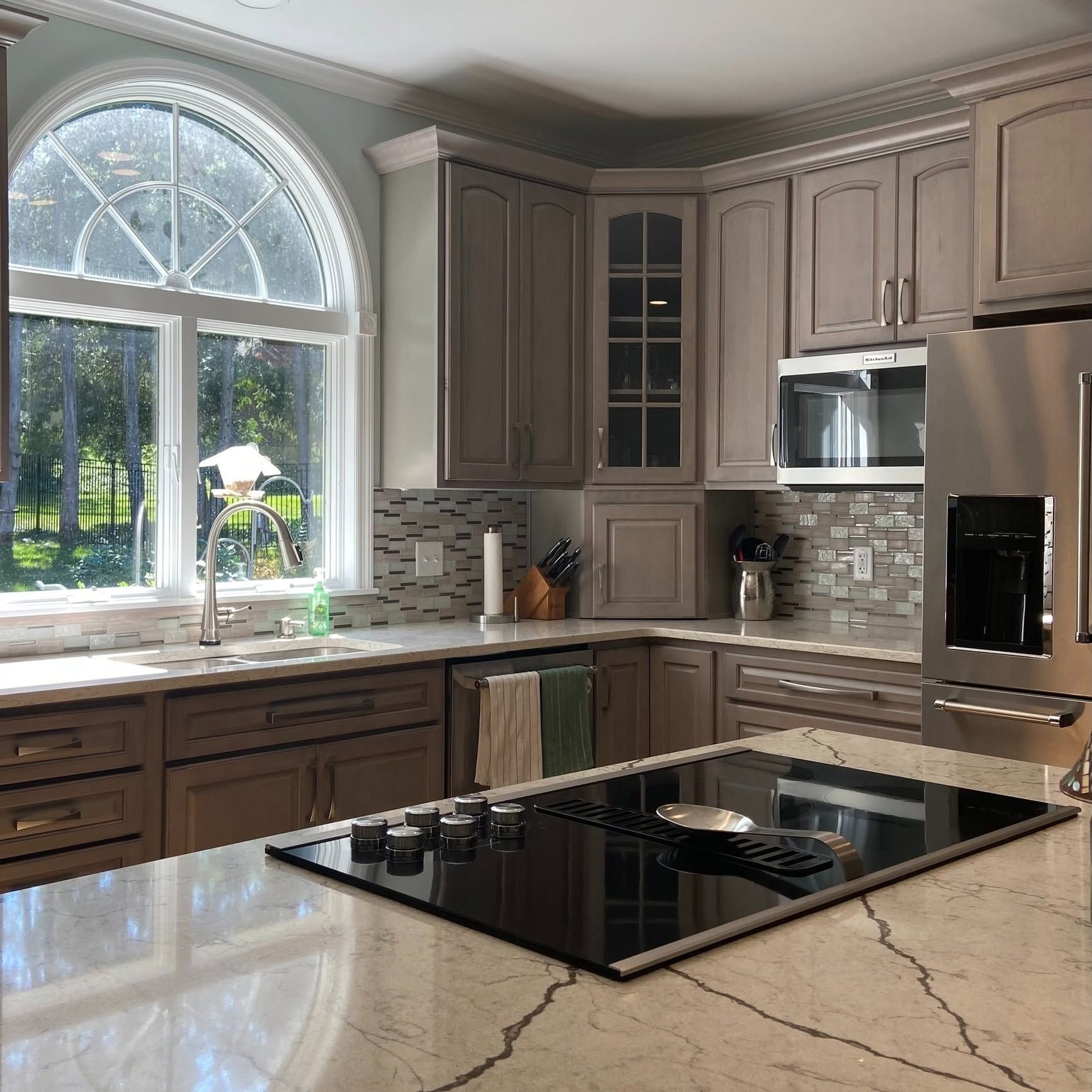 Modern kitchen with gray cabinets, marble countertops, large arched window, and black cooktop island