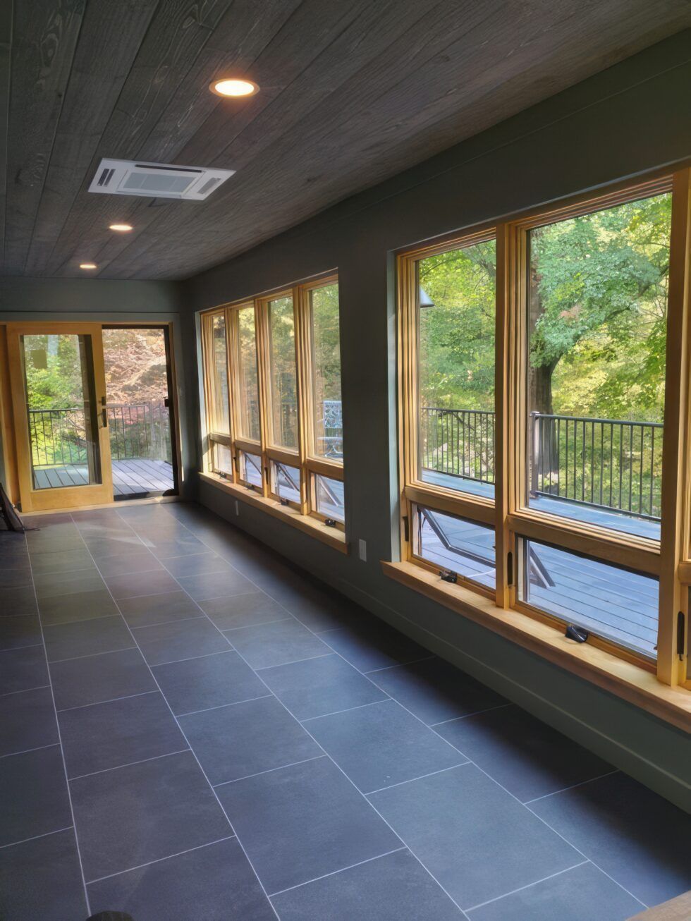 Empty sunroom with dark tile floor, wood ceiling, and large windows overlooking trees and a deck