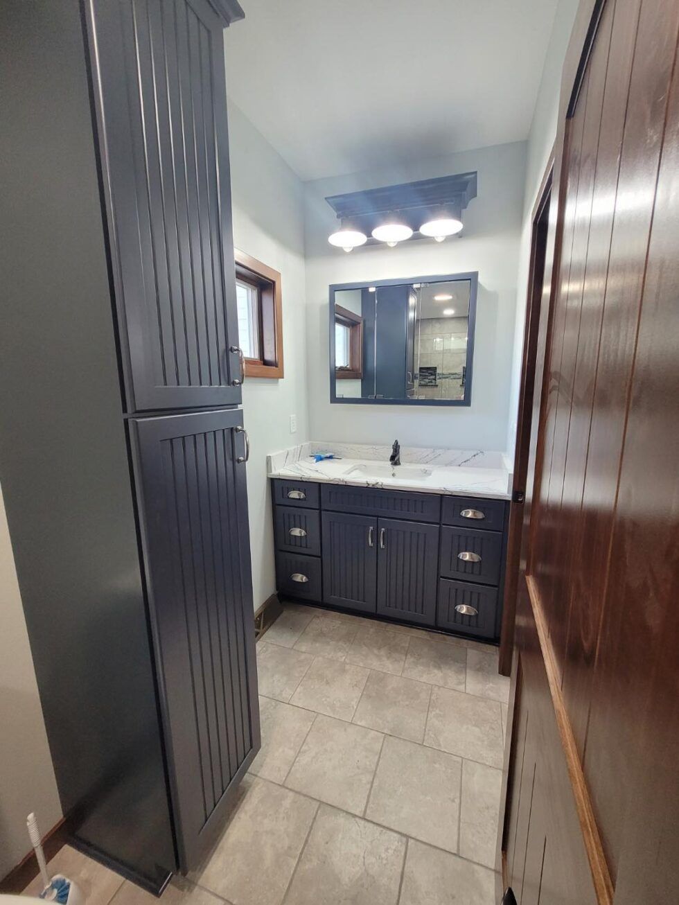 Narrow bathroom with dark vanity, white sink, large mirror, and tiled floor.