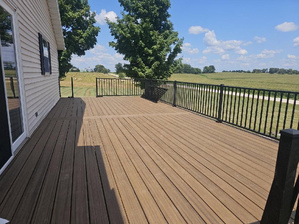 Wooden deck beside a house, with black railing and open field under a blue sky