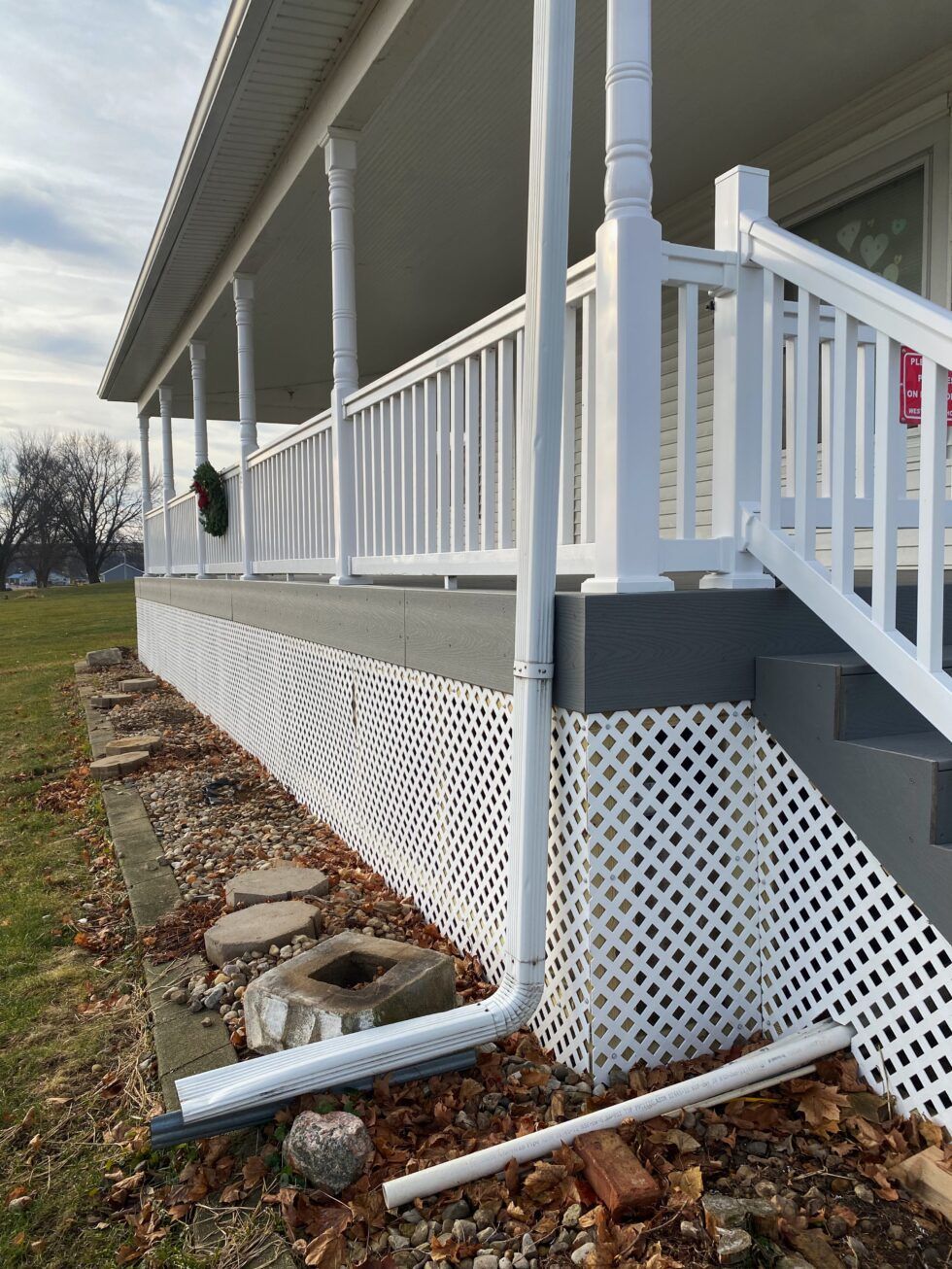 White house porch with lattice skirting and a downspout draining onto a gravel bed.