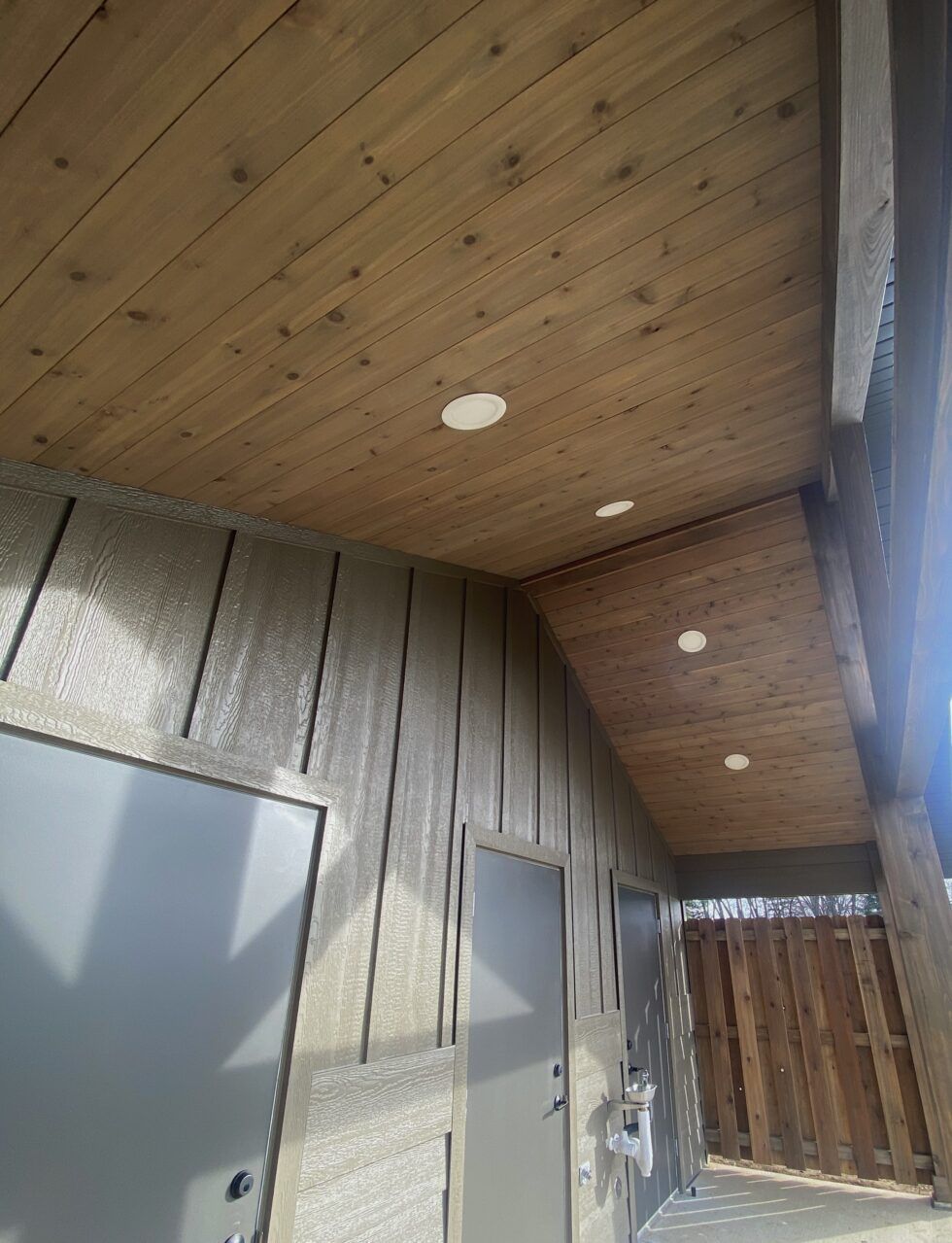 Wood-paneled porch ceiling with recessed lights beside a light gray exterior wall and wooden gate.