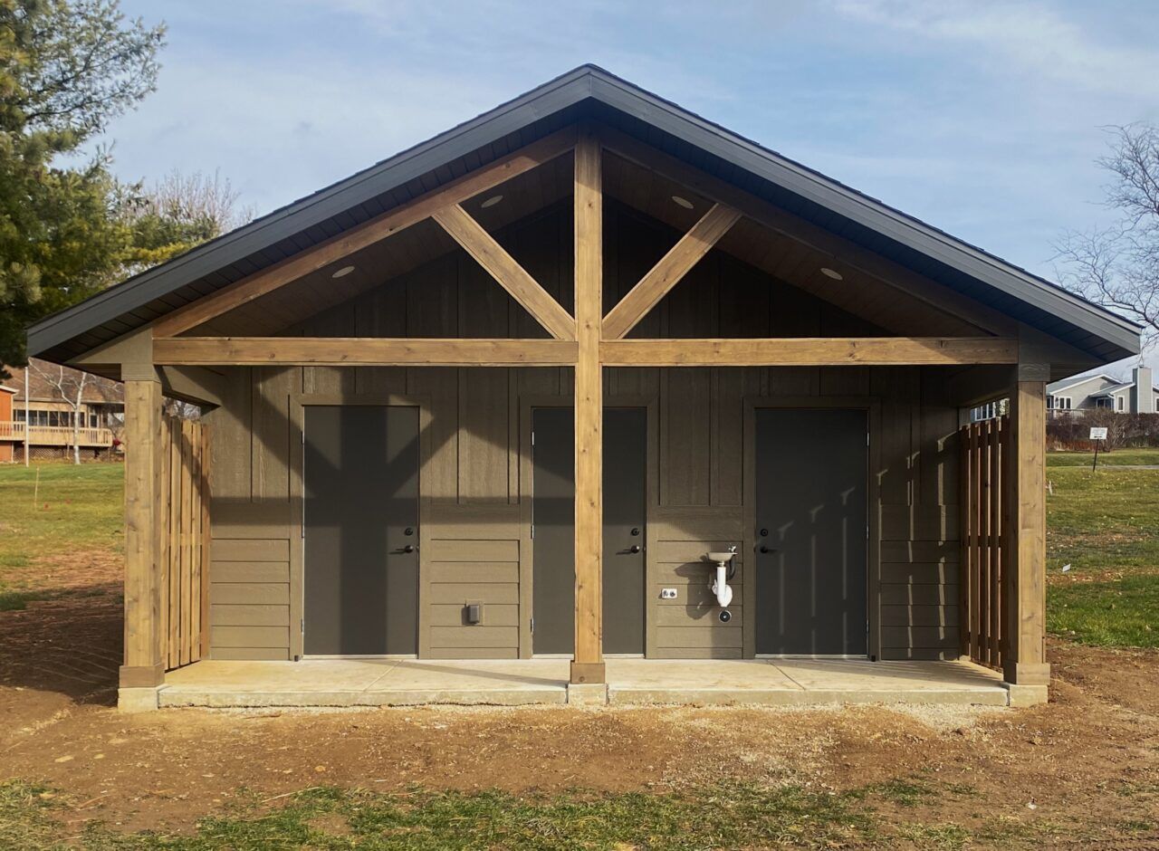 Small tan wooden shed with a peaked roof and open front, showing two doors inside on a grassy lot.