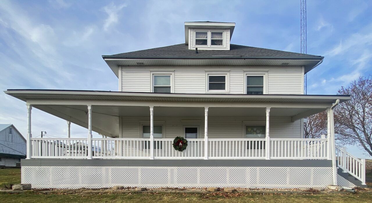 White two-story house with a wraparound porch and dormer windows under a partly cloudy sky
