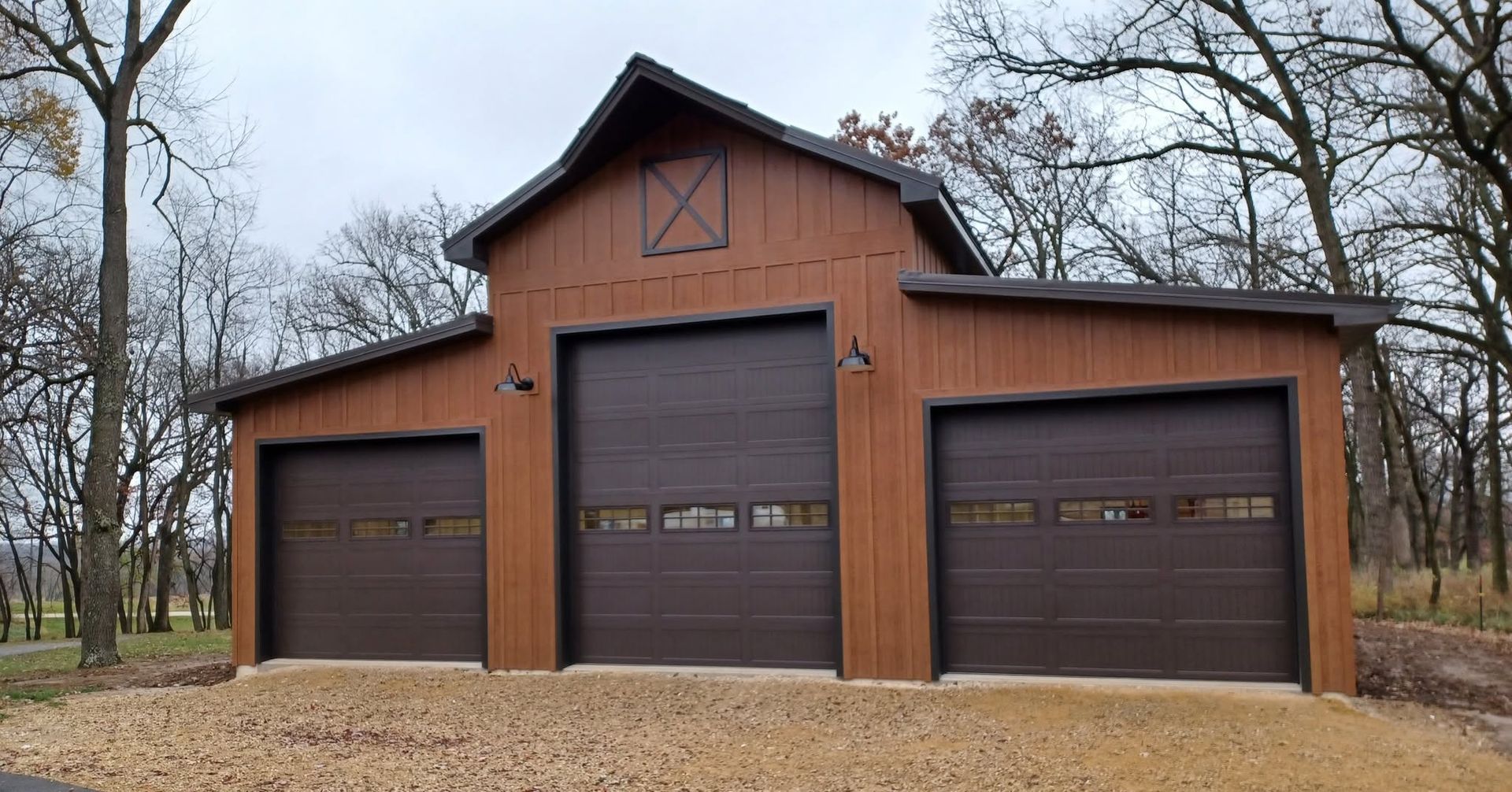 Three-car brown garage with peaked roof and gravel driveway, surrounded by leafless trees