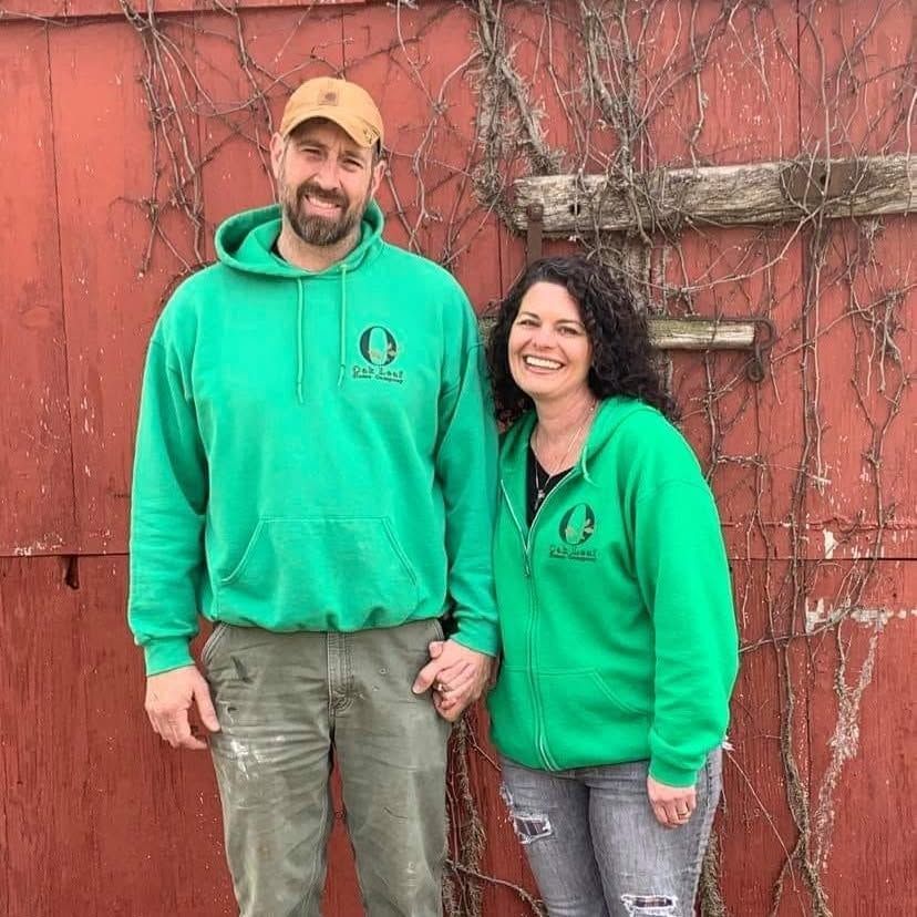 Two smiling people in green hoodies stand against a red wall with bare vines.
