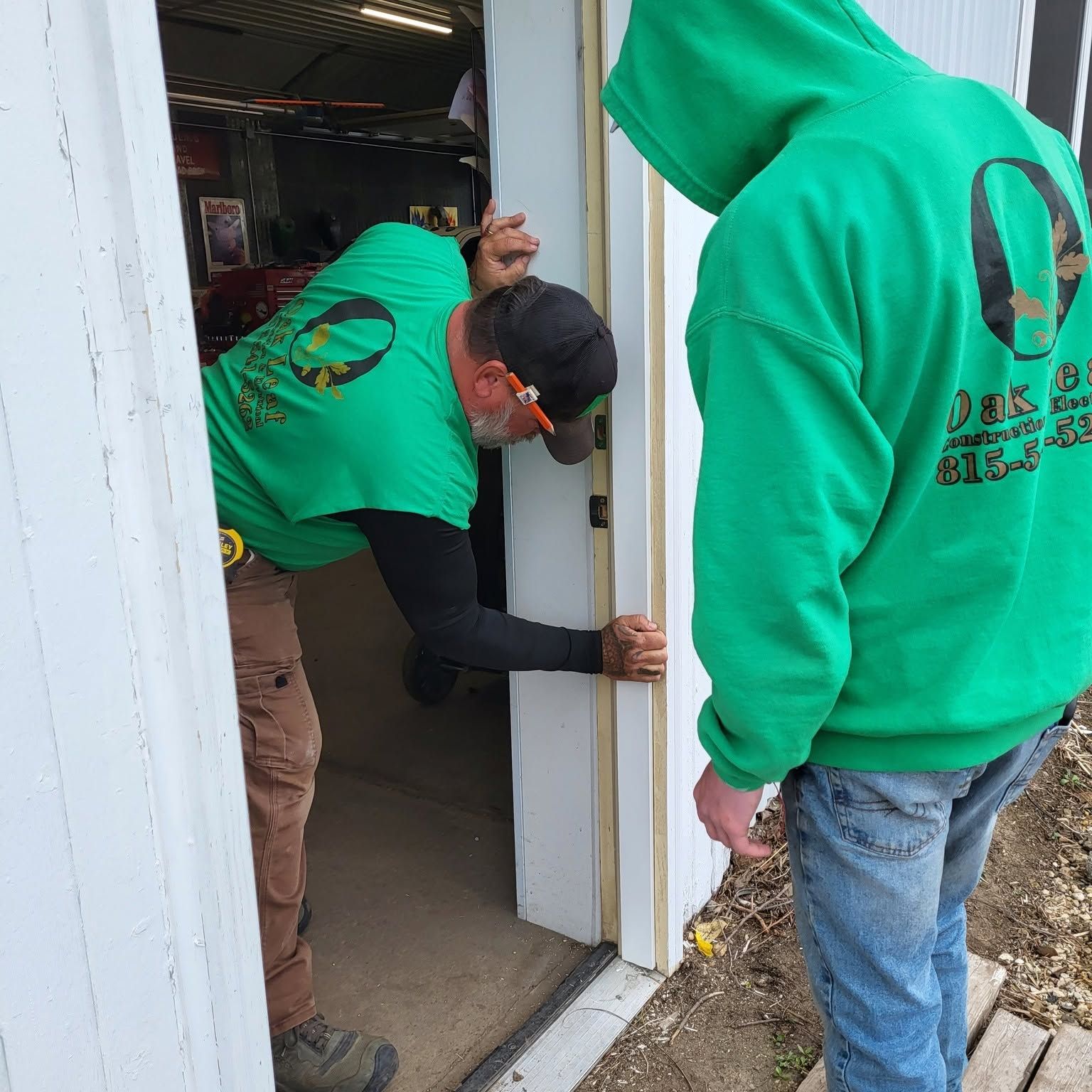 Two workers in green hoodies install a door frame in a small building entrance.