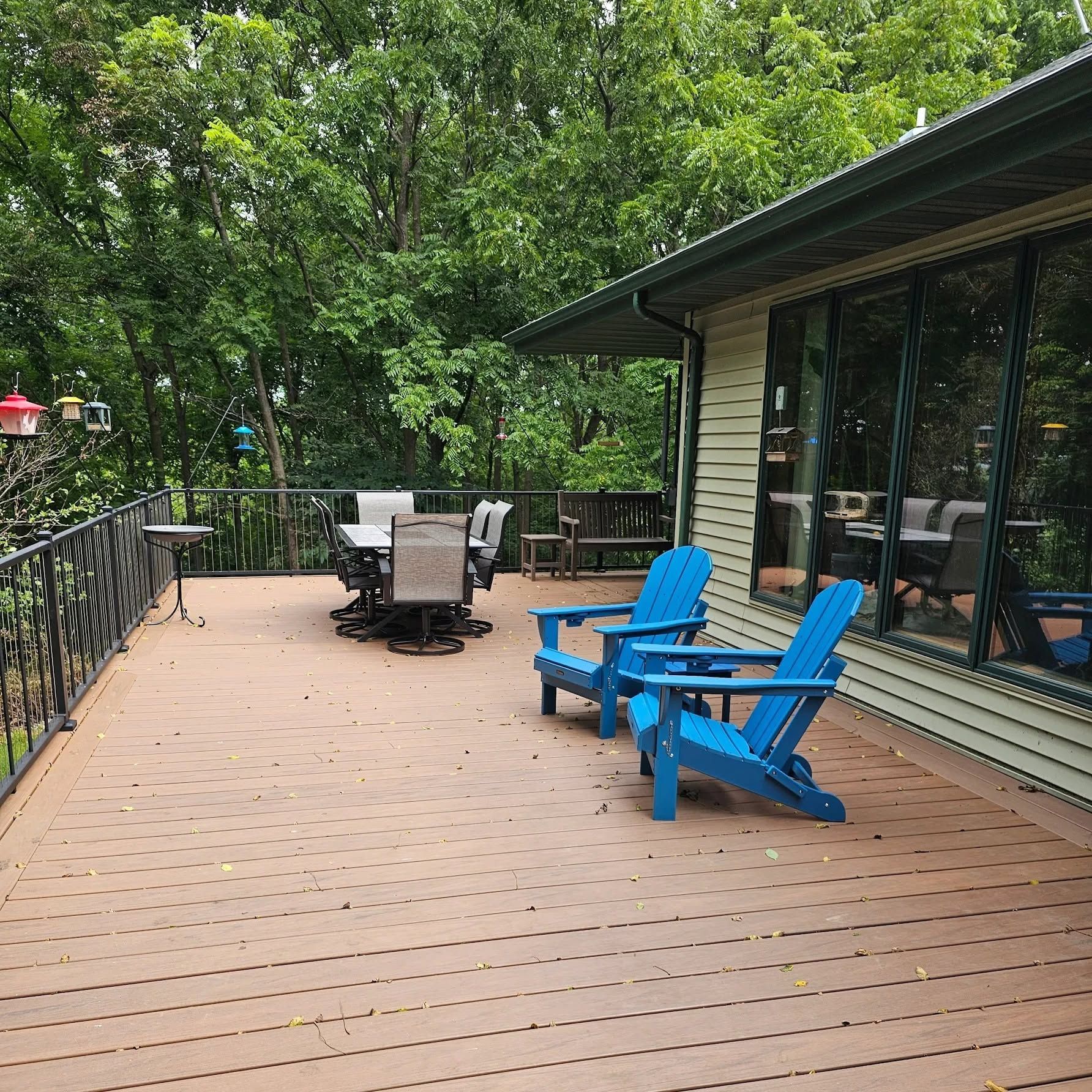 Wooden deck with blue Adirondack chairs beside a house, surrounded by trees and outdoor seating.