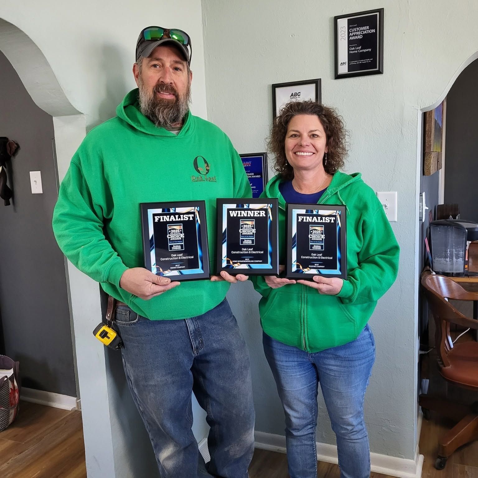 Two people in green hoodies hold framed awards, smiling indoors.