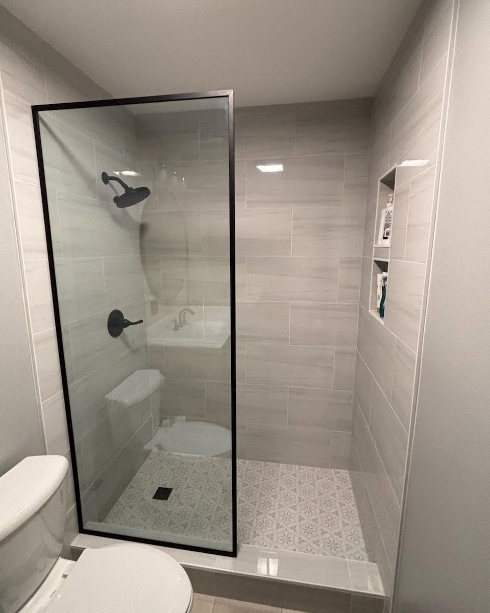 Modern bathroom with glass walk-in shower, gray tile, and white toilet partially visible.