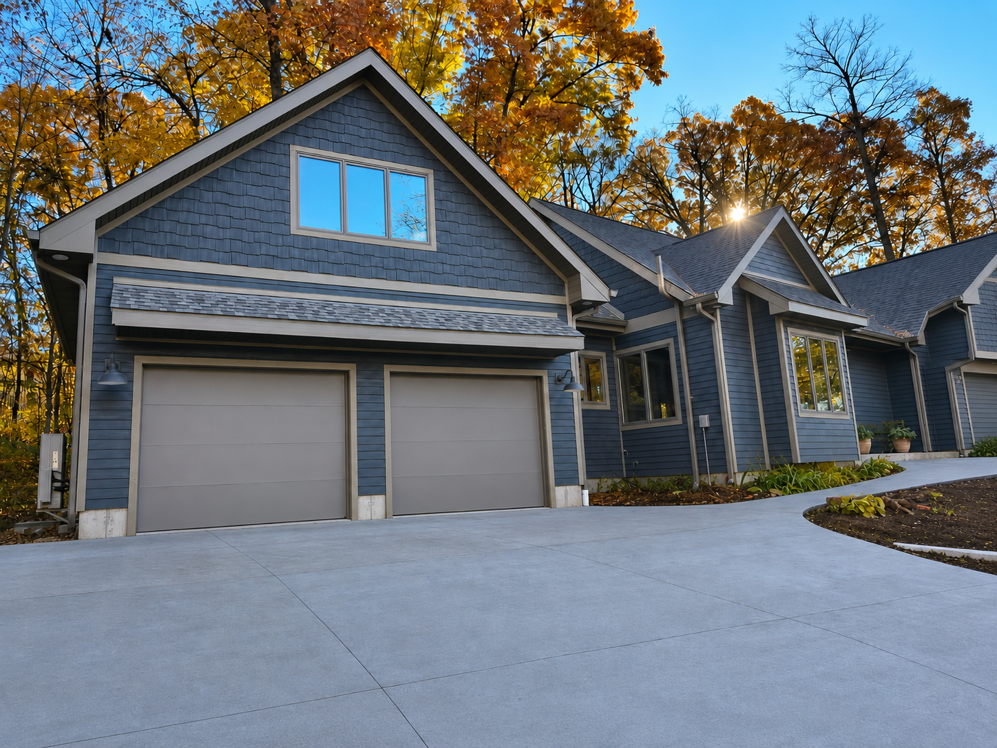 Two-car garage with gray siding and a wide concrete driveway, framed by autumn trees.