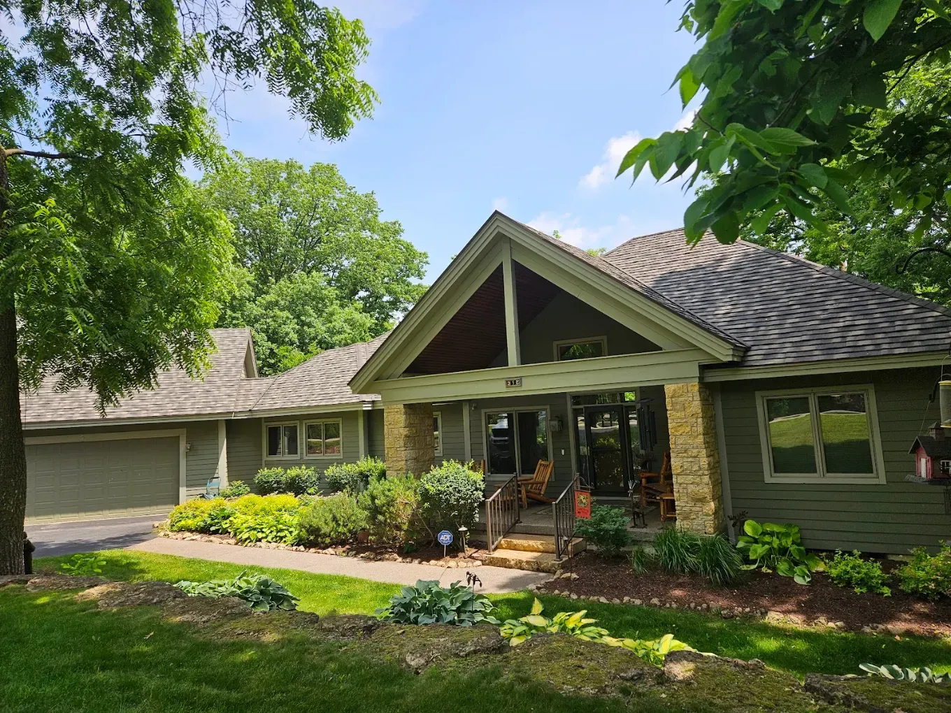 Green house with front porch, white garage, and lush landscaping under trees