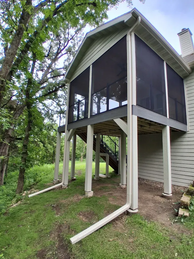 Elevated screened porch on a house, with white posts, stairs, and green yard below.