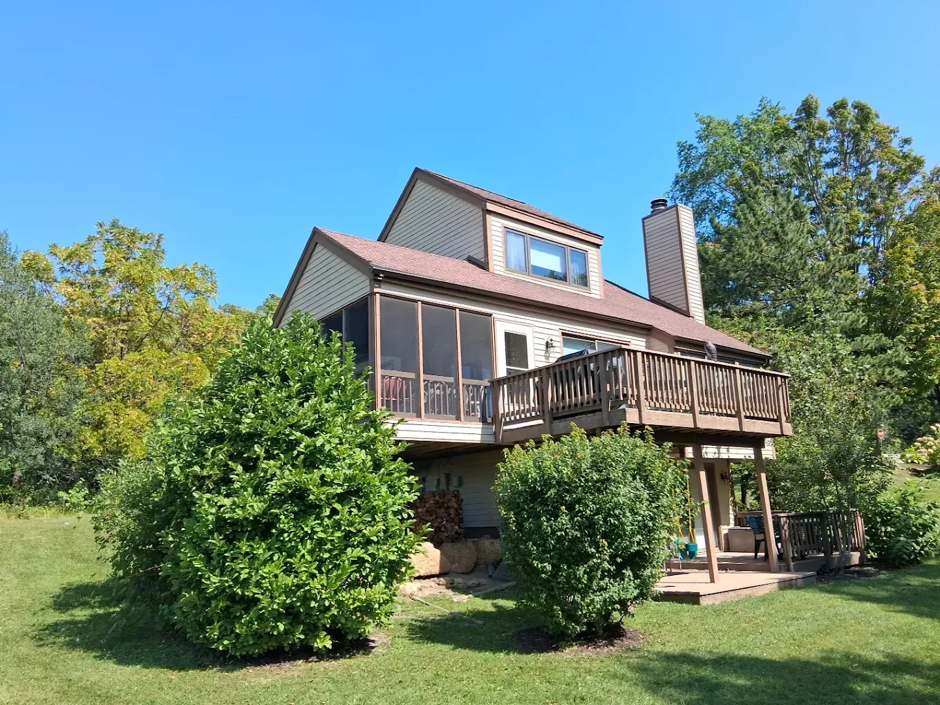 Two-story house with wooden deck and screened porch, surrounded by green trees and shrubs under a blue sky