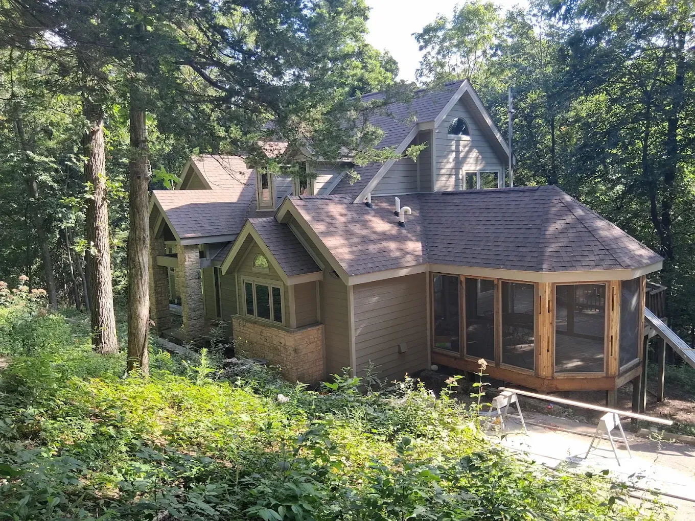 Wooden house with a screened porch in a wooded area, surrounded by trees and greenery
