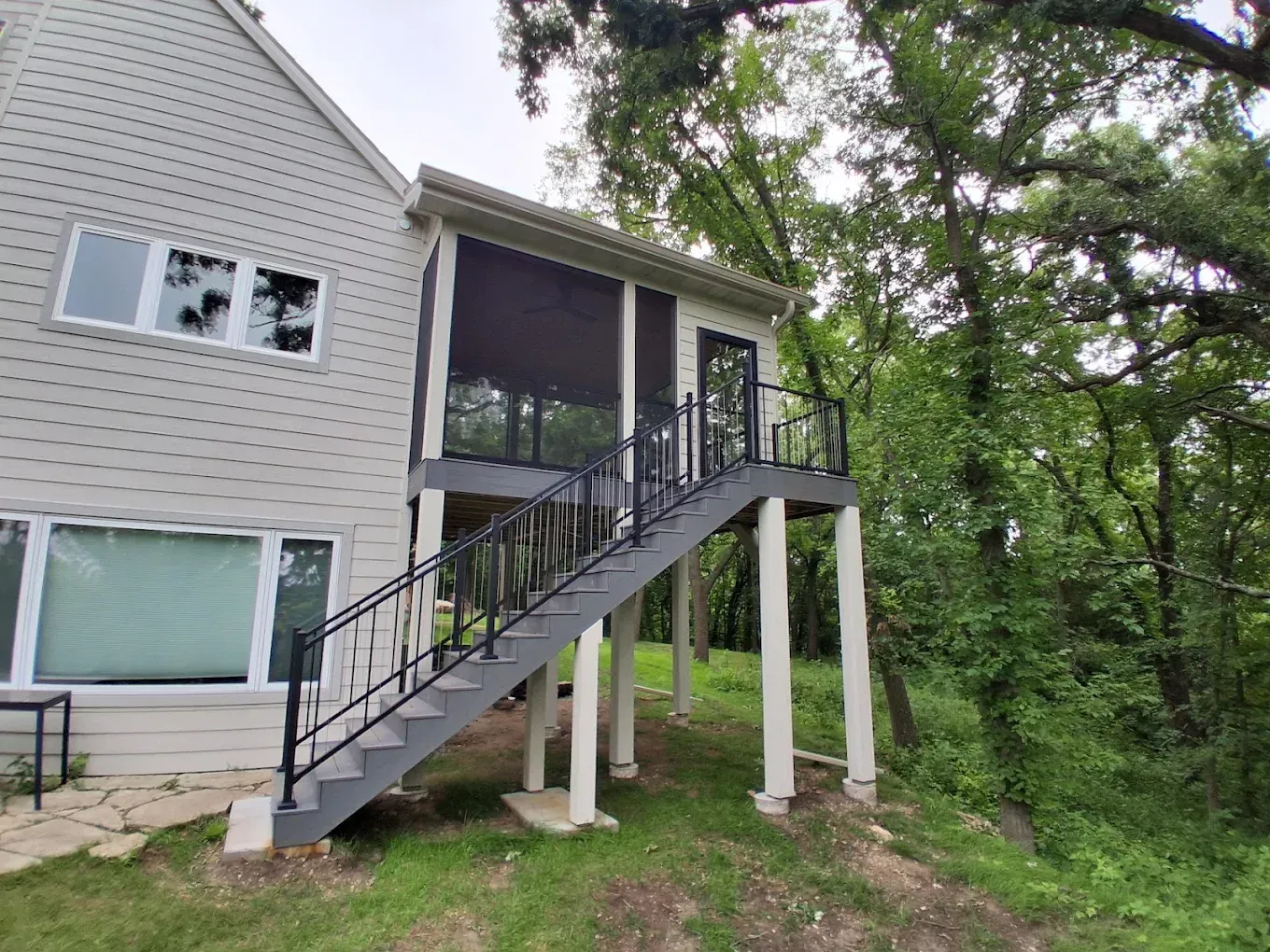Raised porch with exterior stairs on a light gray house beside trees and greenery