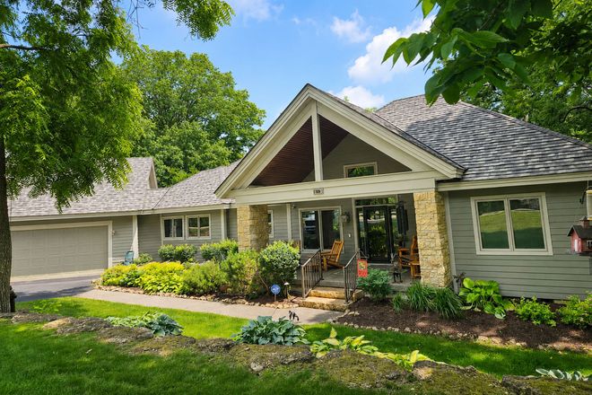 Single-story suburban house with stone entry, gray siding, and lush green trees and lawn