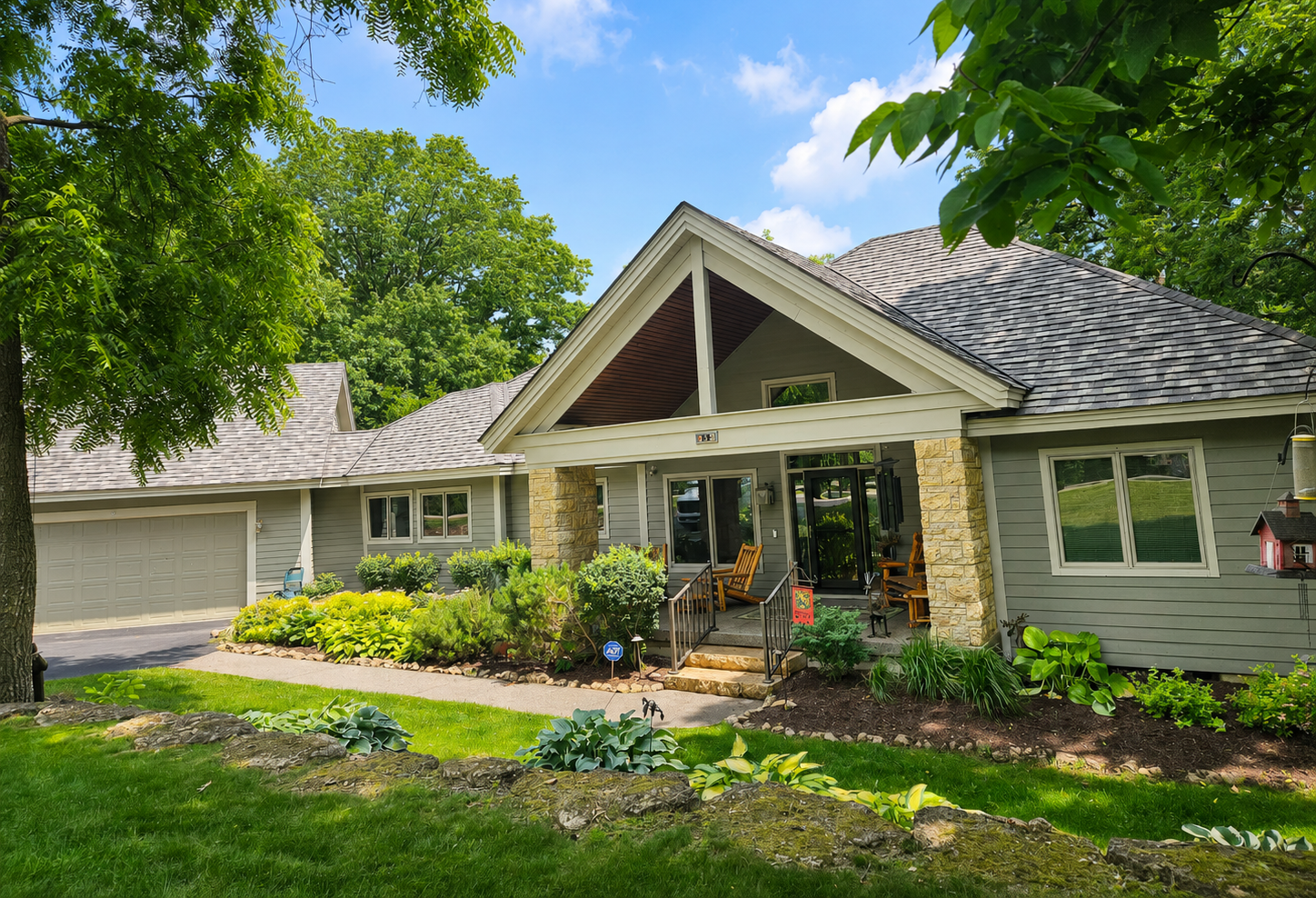 Suburban house with stone porch, gray roof, and green lawn under sunny trees
