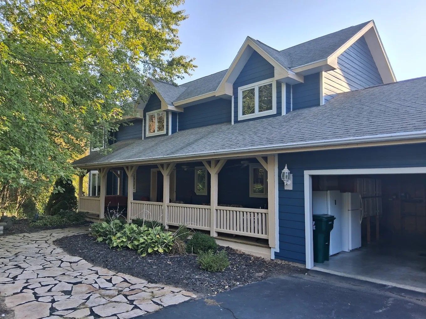 Blue-gray house with a covered porch, dormer windows, and an open garage beside a stone walkway