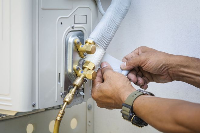 Hands connecting HVAC refrigerant lines to an outdoor unit with brass fittings and white insulation hose