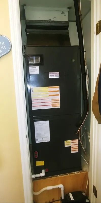 Tall black HVAC furnace unit in a narrow closet with warning labels and visible white piping.