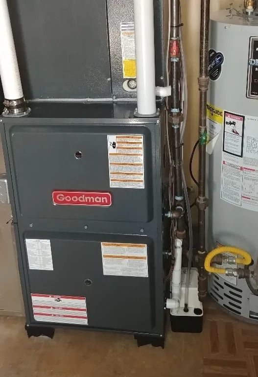 Goodman furnace and water heater in a utility closet with visible pipes and vents