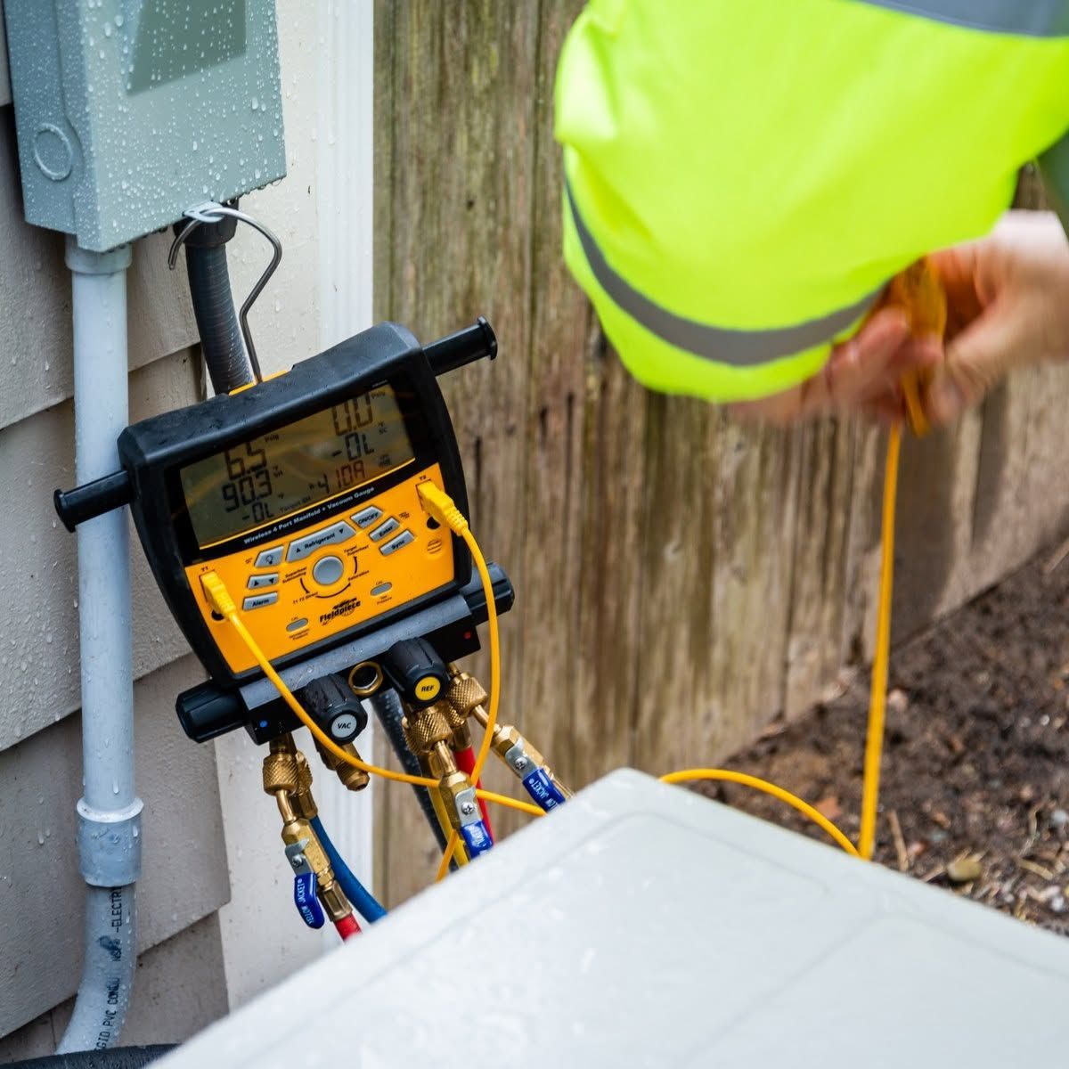 Technician using a yellow HVAC gauge manifold beside an outdoor unit and electrical panel