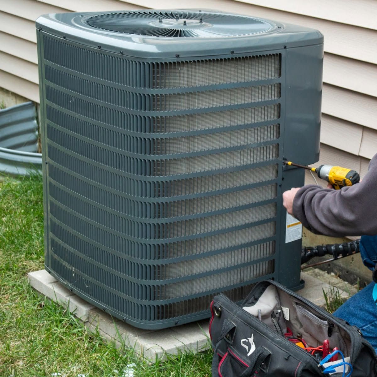 Technician using a drill to service an outdoor air conditioner unit beside a house