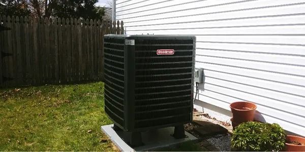 Outdoor air conditioning unit beside a house, with a small potted plant nearby and a fenced yard.