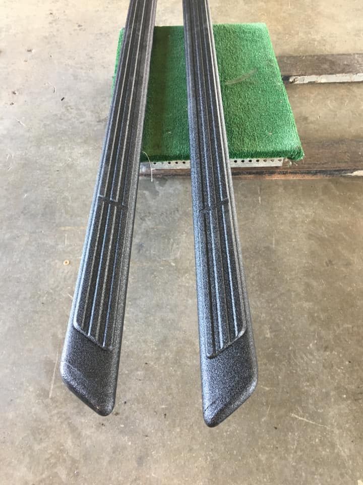 Pair of black, textured running boards, resting on a green surface, for a vehicle.