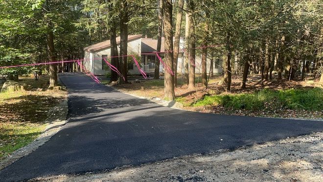 Asphalt driveway leading to a stone house surrounded by trees.