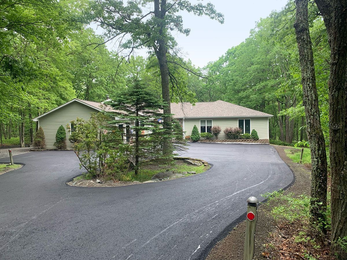 Asphalt driveway curves toward a beige house surrounded by trees.