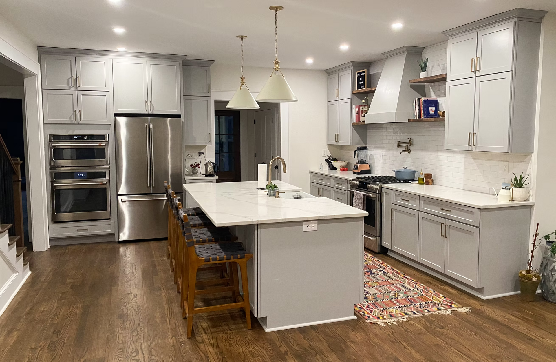 A modern kitchen with gray cabinets, stainless steel appliances, and a large island with seating. Hardwood floors, white countertops, and pendant lights.