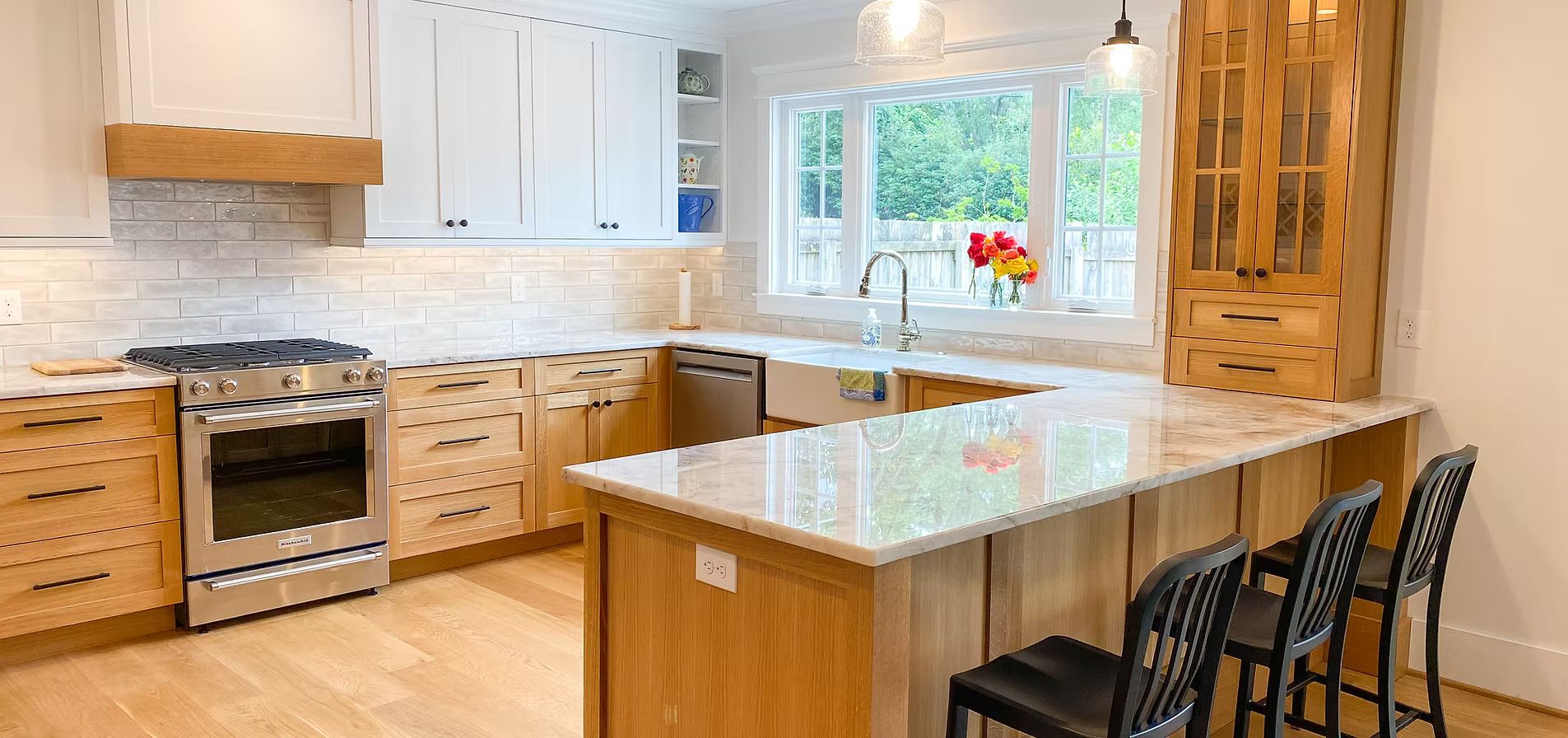 A modern kitchen with light wood cabinets, white countertops, and a stainless steel range. A breakfast bar has three dark chairs.