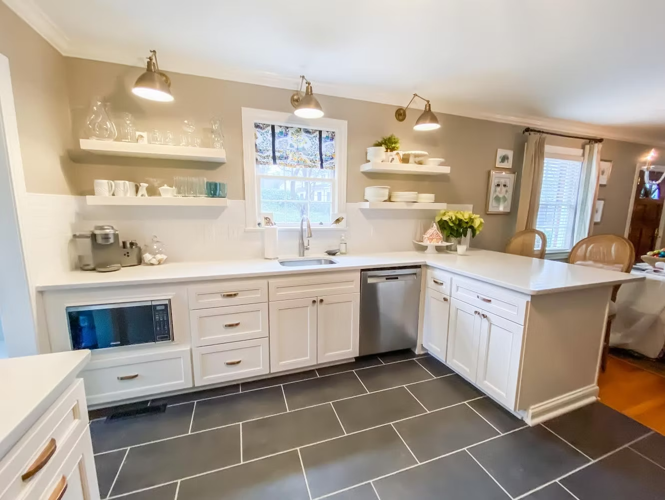 A white kitchen with gray tile floors, featuring white cabinets, countertops, and floating shelves. Stainless steel appliances and a kitchen island are also present.